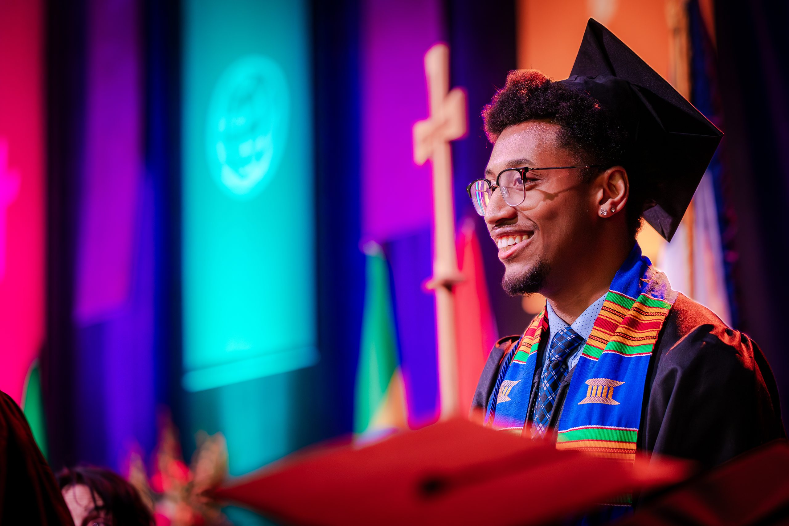 A graduate in a cap and gown smiles broadly during a ceremony, with colorful stage lighting and a cross visible in the background.